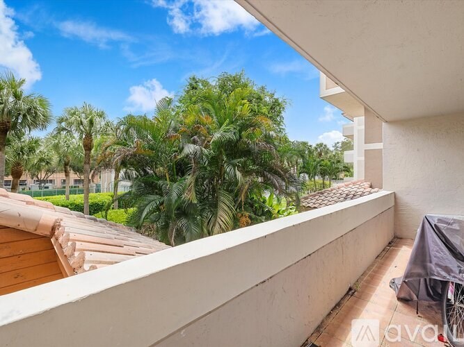 A balcony with a white wall and a bicycle is overlooking a garden with palm trees.