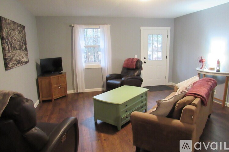 A living room with a black leather couch, a green ottoman, a wooden cabinet, a flat screen TV, a window with white curtains, a white door, and a painting on the wall.