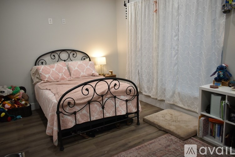 A bed with a pink blanket and a black metal headboard is in the corner of a room with a white curtain.