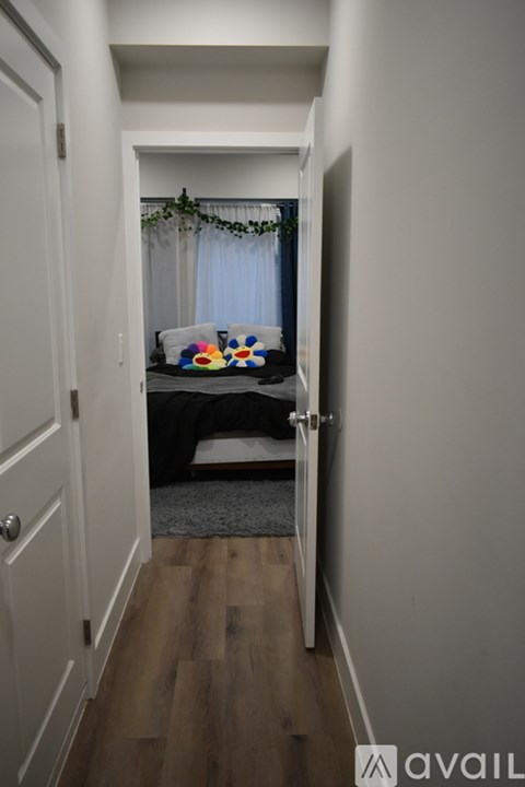 A hallway with a door on the left and a bedroom with a bed and a window on the right.