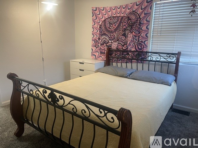 A bed with a white sheet and a pillow is in the foreground of a room with a patterned wall hanging.