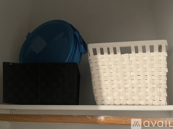 A white wicker basket sits on a shelf next to a black box and a blue object.