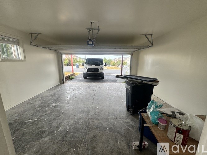 A white van is parked in a garage with a trash bin and some paint cans on the floor.