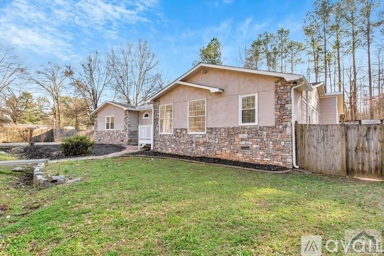 A house with a stone wall and a white door is surrounded by a grassy area and trees.