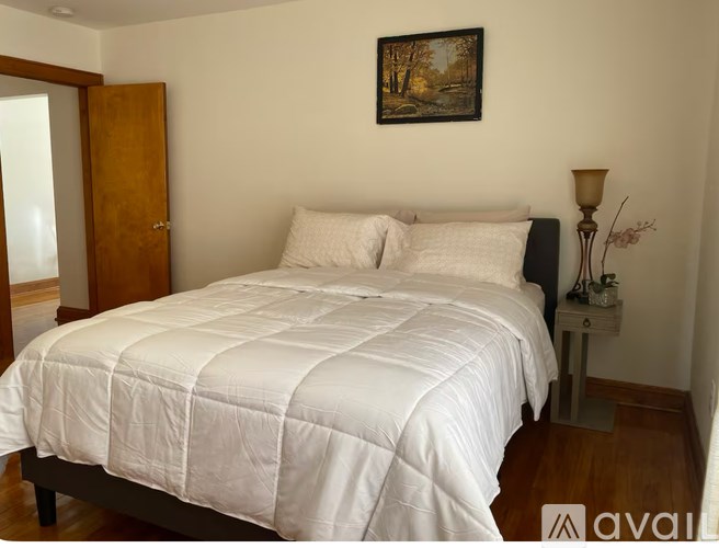 A bed with a white comforter and two pillows sits in a room with a painting on the wall and a small table with a lamp.