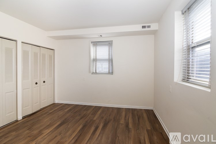 A room with wooden floors and white walls, featuring a window with blinds and a small air vent on the wall.