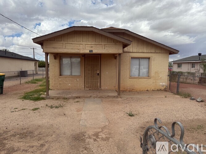 A small, single-story house with a brown roof and a brown door is surrounded by a dirt lot.