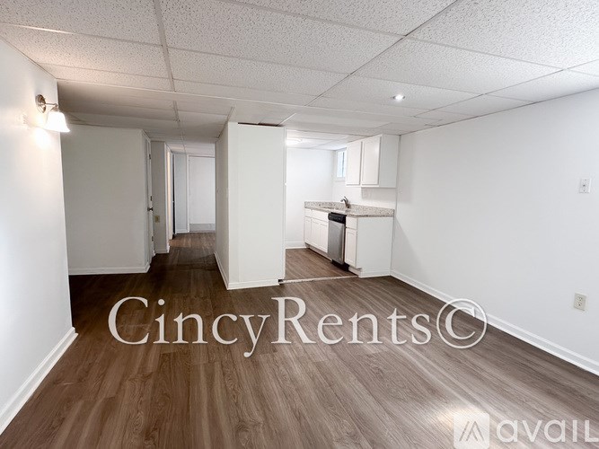 A well-lit, empty room with wooden flooring and white walls, with a kitchen area visible in the background.