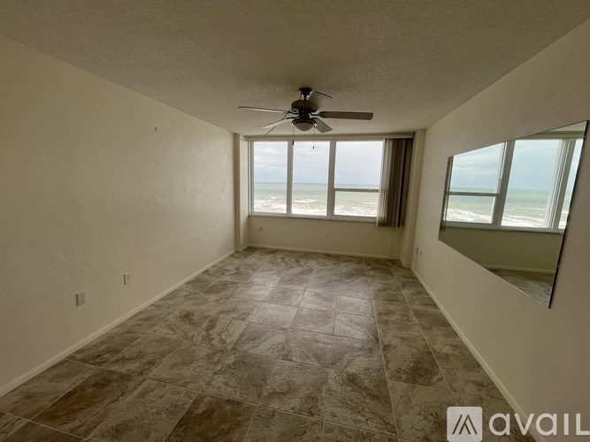 A room with a ceiling fan and a view of the beach through the windows.