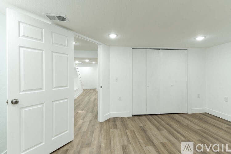 A white door is on the left of a room with a wooden floor and white walls.