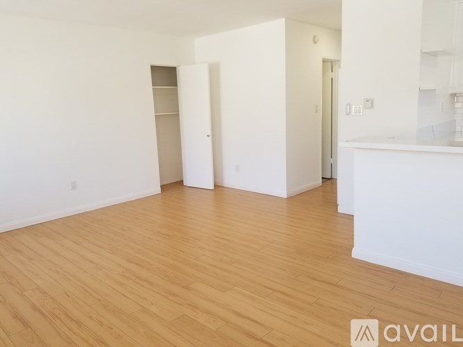 A room with wooden flooring and white walls, with a kitchen area to the right.