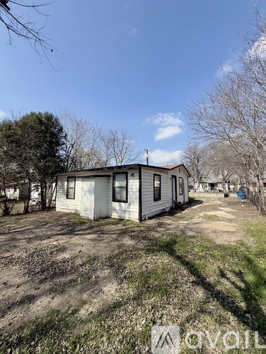 A small white house with a brown roof is surrounded by a grassy area and leafless trees.