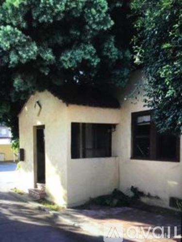 A small white house with a black door and windows is surrounded by trees.