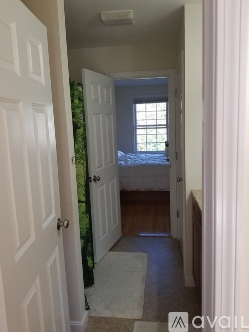 A hallway with a door on the left and a bedroom with a bed and window on the right.