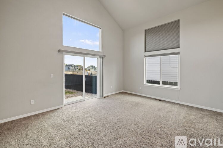A room with a carpeted floor, a window with blinds, and a view of a fence and houses outside.