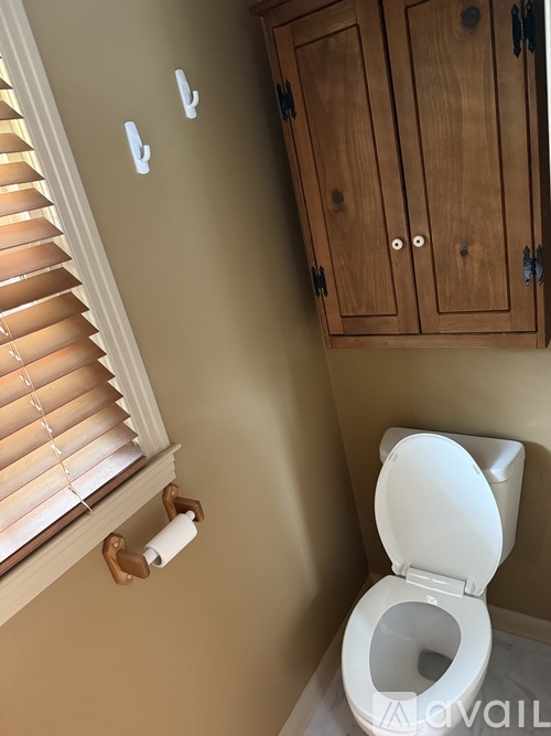 A toilet in a small bathroom with a wooden cabinet and a window with blinds.