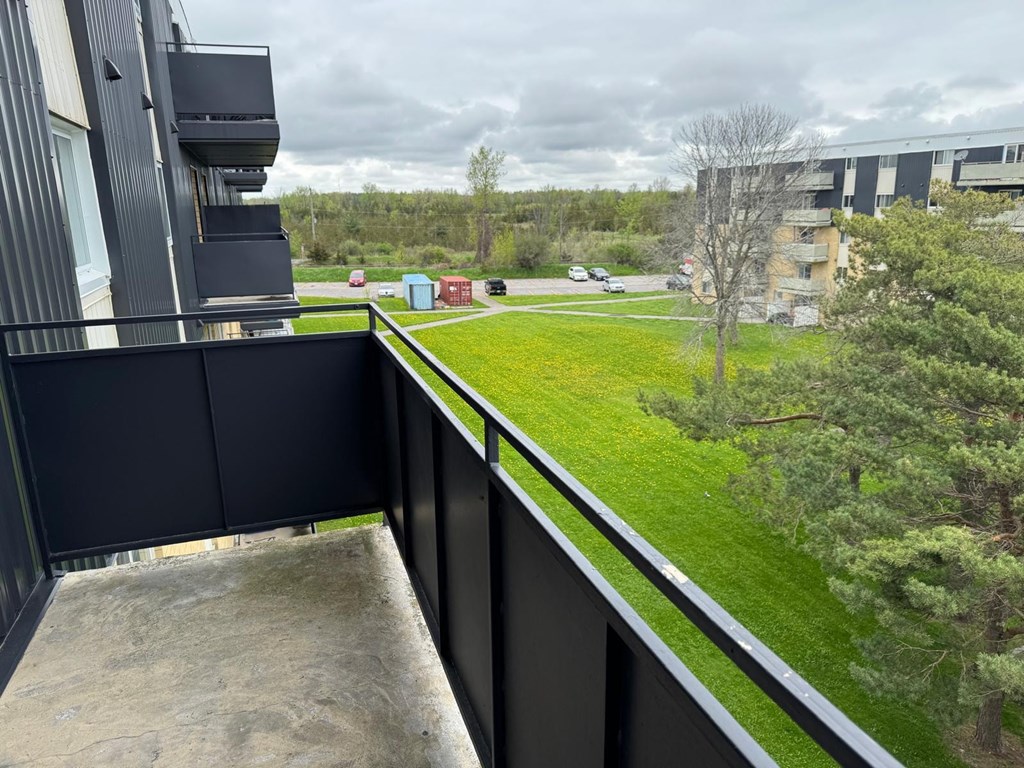 A balcony with a black railing overlooks a grassy area with trees and a building in the distance.