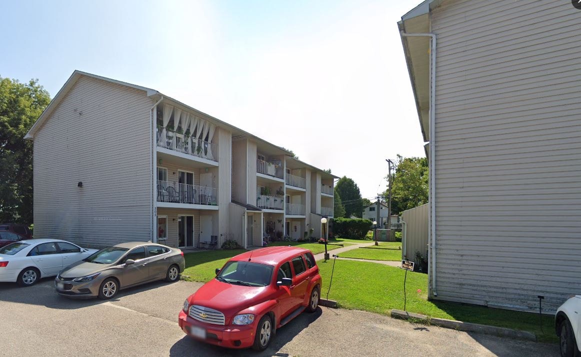 A red car is parked in front of a white building with a balcony.