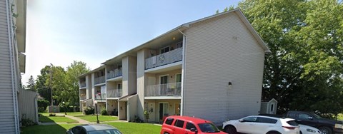 A red car is parked in front of a white building with a balcony.