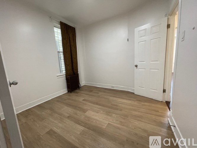 A room with wooden flooring and white walls, with a door on the left and a window with brown curtains.