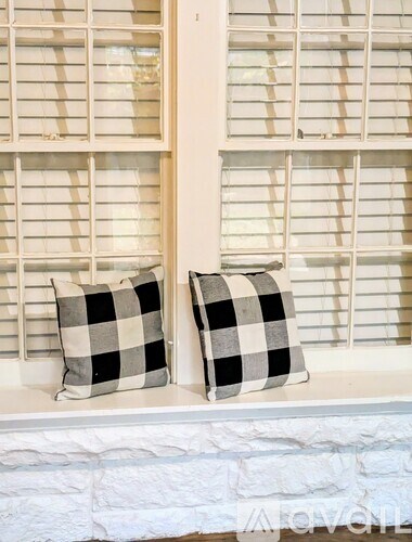 A room with a wooden floor, a window with blinds, a white door with a shelf above it, and a wall with a switch.