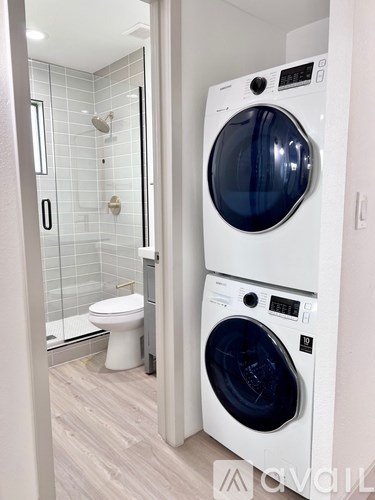 A white washing machine is in a bathroom with a toilet and a shower.