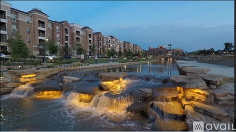 A waterfall in a park with buildings in the background.
