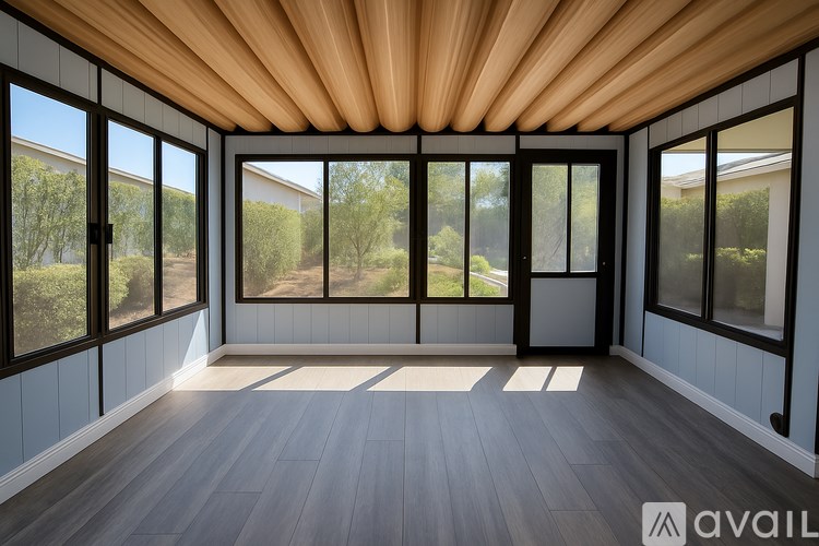 A room with a wooden ceiling and floor, large windows, and a view of trees outside.