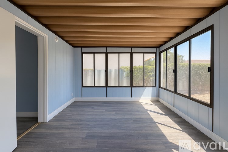 A room with a wooden ceiling and floor, white walls, and a view of a fence and trees outside through the windows.