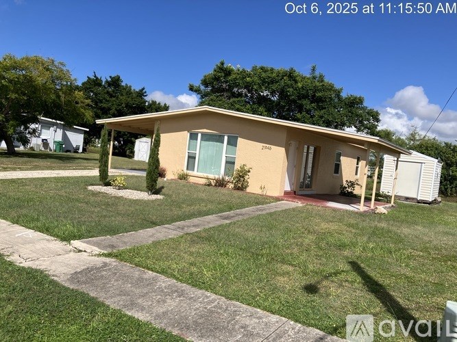 A house with a yellow wall and a red door is surrounded by a grassy lawn and a concrete pathway.