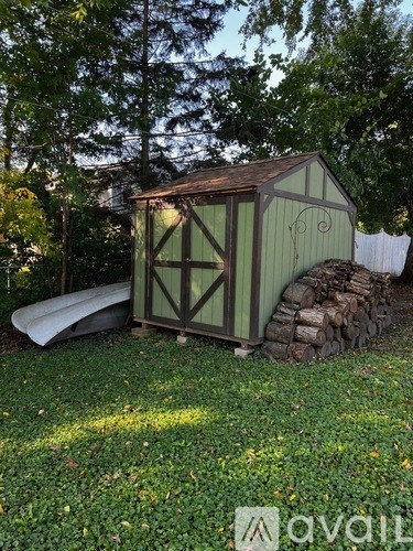 A shed with a green roof and white walls is surrounded by a pile of wood and a white fence.