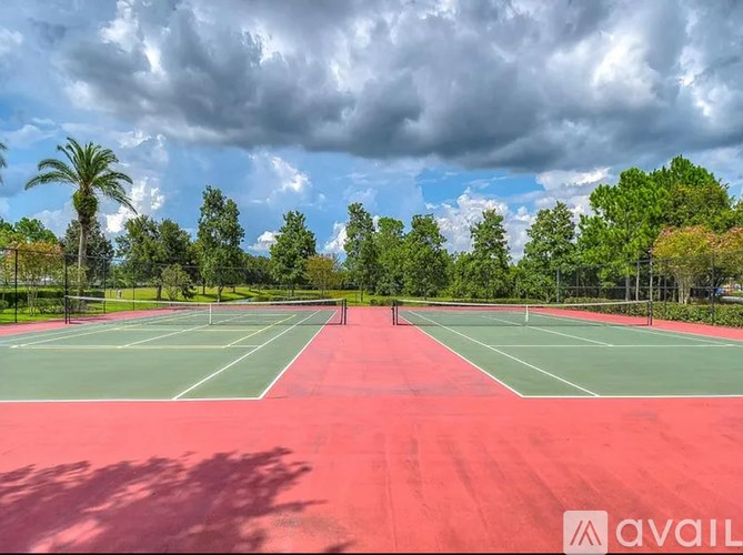 A tennis court with a red surface and white lines, surrounded by trees and a cloudy sky.