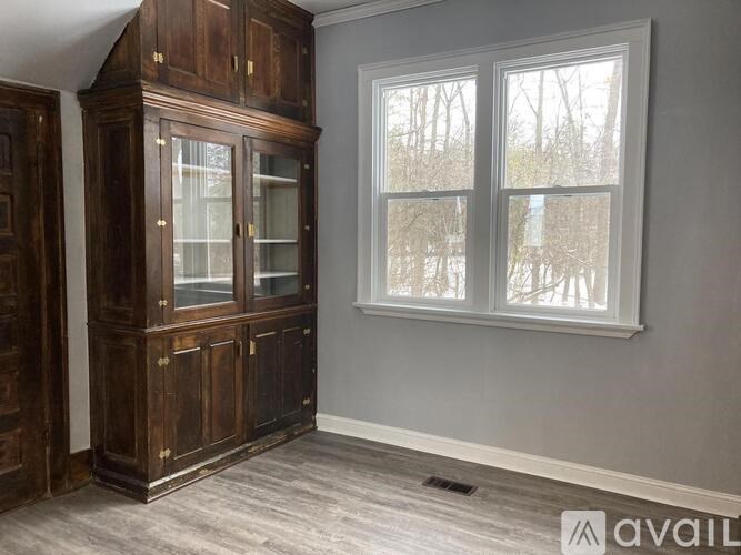 A large wooden cabinet with glass doors sits in a room with a window showing trees outside.