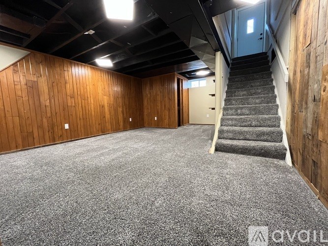 A room with wooden walls and a carpeted floor with a staircase on the right side.