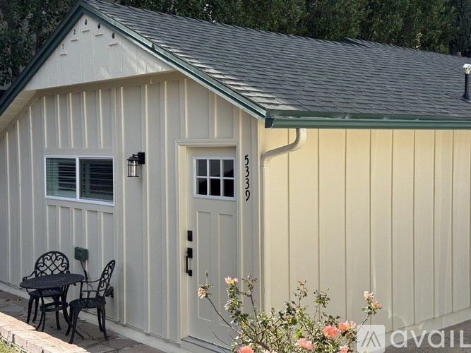 A small house with a white door and windows is surrounded by flowers.