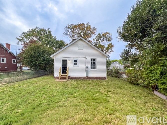 A small white house with a brown door and a window is surrounded by a grassy area and trees.