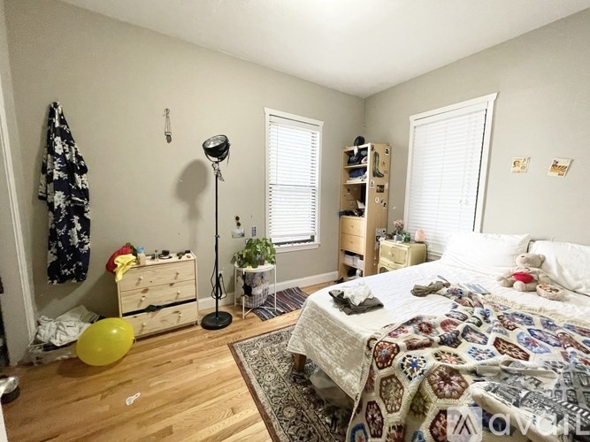 A bedroom with a bed, a dresser, a nightstand, a window with blinds, a lamp, a plant, and a rug.