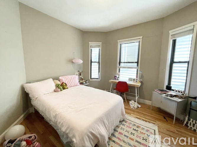 A bedroom with a white bed, a desk with a chair, and a window with blinds.