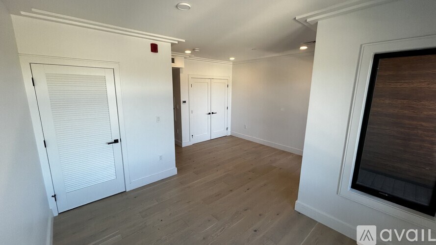 A room with white walls and wooden flooring, featuring a large window with blinds and a door with a handle.
