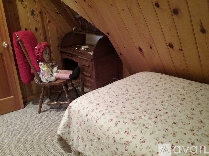 A doll is sitting in a rocking chair in a room with wooden walls and a bed with a floral comforter.