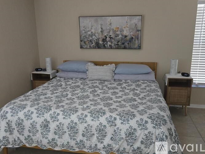 A bed with a floral patterned comforter and two pillows is in the foreground with a painting on the wall above it.