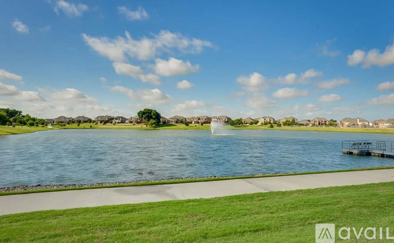 A serene lake with a fountain in the middle, surrounded by a grassy area and buildings in the distance under a clear blue sky.