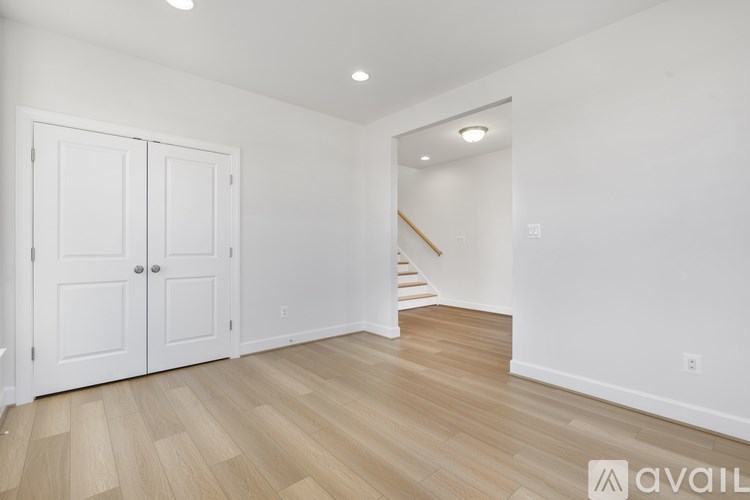 A room with white walls and wooden flooring, with a white door on the left and a staircase in the background.