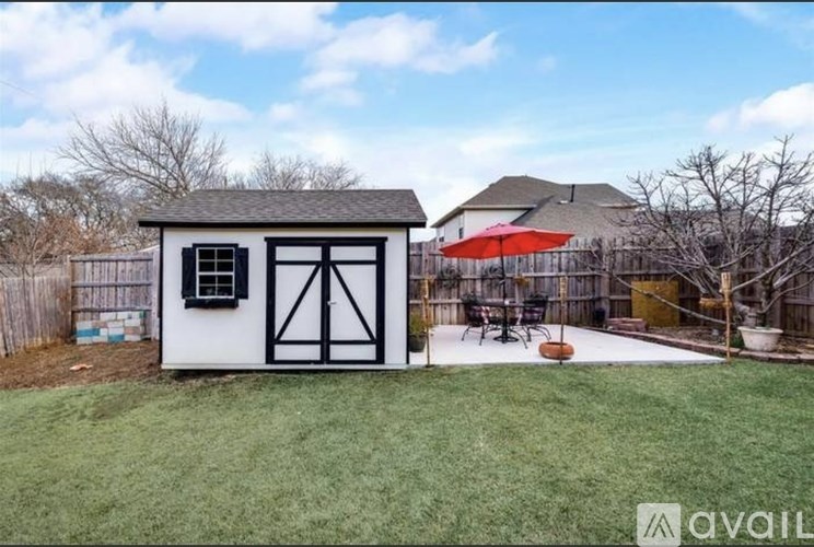 A small white building with a black door and window is situated in a grassy area with a patio and umbrella in the background.