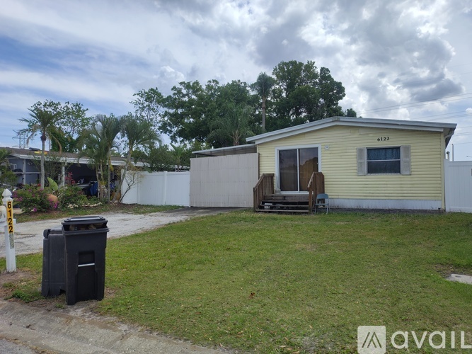 A small house with a yellow exterior is surrounded by a grassy area and a trash can is placed on the left side of the image.