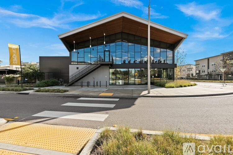 A modern building with a glass facade and a wooden roof is surrounded by a grassy area and a street with a pedestrian crossing.