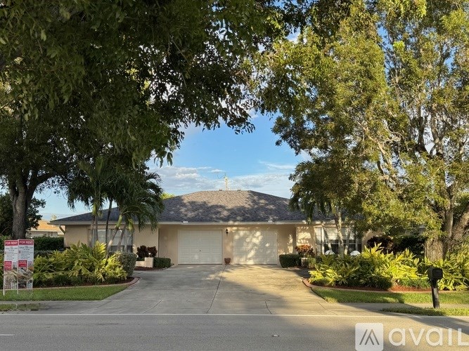 A house with a garage is surrounded by greenery and has a sign in front.