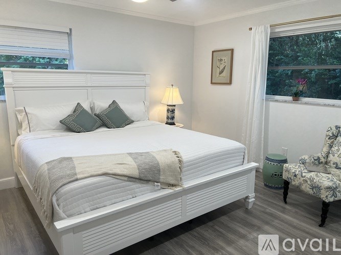 A white bed with a grey and white striped blanket is in a room with a lamp and a chair.
