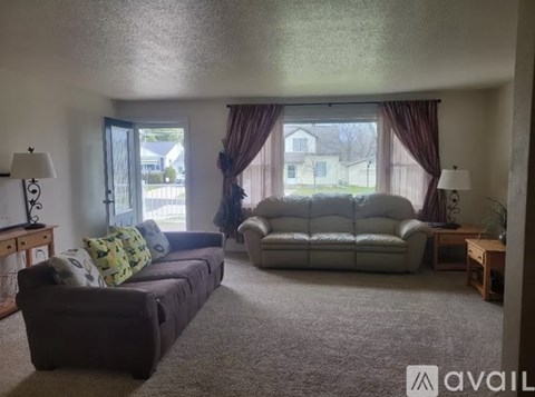 A living room with a grey carpet and a large window with curtains.