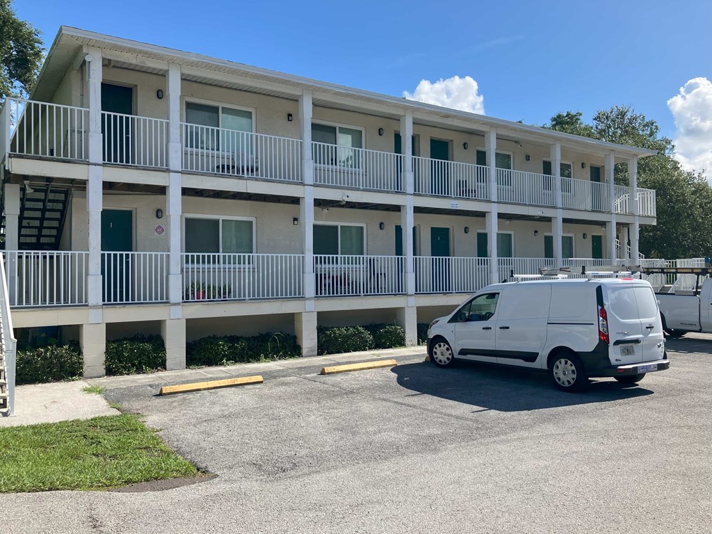 A white van is parked in a parking lot in front of a white building with balconies.